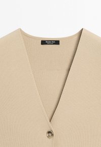 Beiger gestrickter Cardigan mit tiefem V-Ausschnitt und Einzelknopfverschluss, mit einem schwarzen Etikett mit der Aufschrift Massimo Dutti, hergestellt in China.