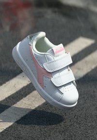 White sneaker with pink accents, Velcro straps, perforated detail on the sides, and a textured pattern on the heel. Soft lining visible.
