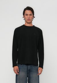 Svart langermet skjorte med rund hals og en subtil merkepute på venstre side, kombinert med falmede blå jeans. Glatt tekstur.