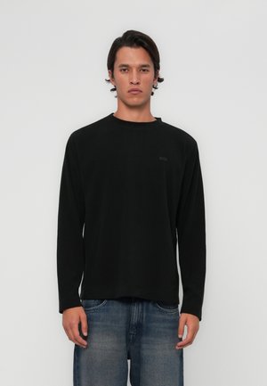 Black long-sleeve shirt with a round neckline and a subtle brand patch on the left side, paired with faded blue jeans. Smooth texture.