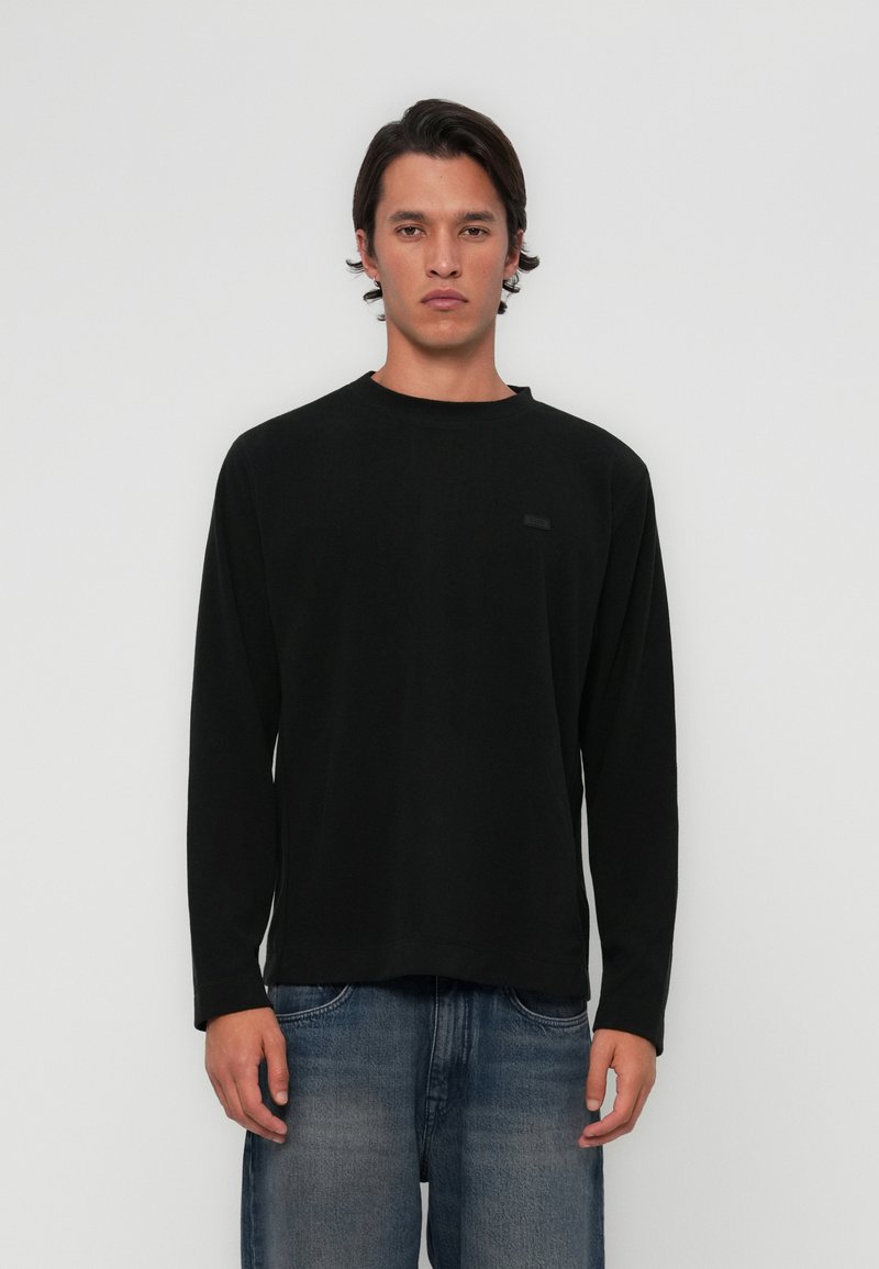 Svart langermet skjorte med rund hals og en subtil merkepute på venstre side, kombinert med falmede blå jeans. Glatt tekstur.