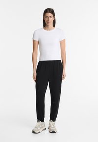 White fitted t-shirt with short sleeves, paired with black elastic waist trousers. Model wears beige and gray athletic shoes.
