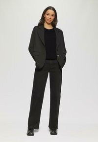 Blazer noir ajusté avec revers en pointe, associé à un top en tricot noir et un jean large noir, complétant le look avec des chaussures noires à clous.