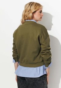 Sweatshirt vert olive avec des poignets et un ourlet côtelés, superposé sur une chemise rayée bleu clair. Tissu doux avec une coupe décontractée.
