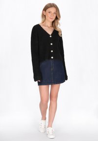 Cardigan corto nero con scollo a V, dotato di cinque grandi bottoni, abbinato a una gonna di jeans e sneaker bianche. Materiale morbido e testurizzato.