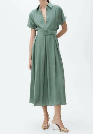 Femme portant une robe midi ceinturée vert sauge avec des manches courtes et des sandales à talons hauts dorées, debout devant un fond uni.