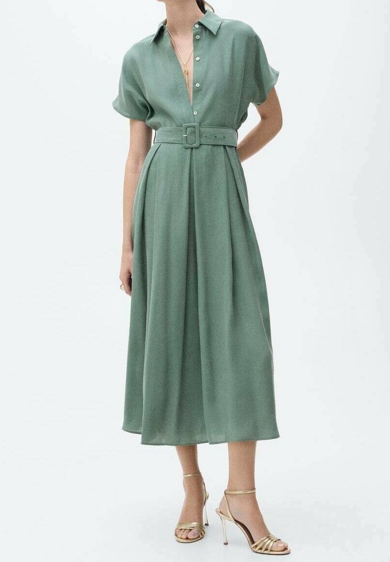 Femme portant une robe midi ceinturée vert sauge avec des manches courtes et des sandales à talons hauts dorées, debout devant un fond uni.