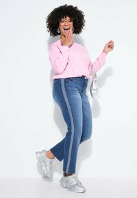 Donna con capelli ricci che ride, indossa una felpa rosa, jeans blu con strisce laterali, sneakers argento, occhiali, orecchini a cerchio e tiene una borsa argento.