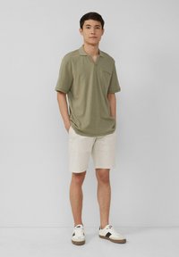 Młody mężczyzna stoi z rękami w kieszeniach, ubrany w oliwkową koszulkę polo, beżowe szorty i białe sneakersy na jednolitym szarym tle.