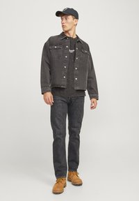 Chaqueta de mezclilla negra con dos bolsillos en el pecho, combinada con una camiseta gráfica negra y jeans oscuros. Botas de trabajo beige complementan el atuendo.