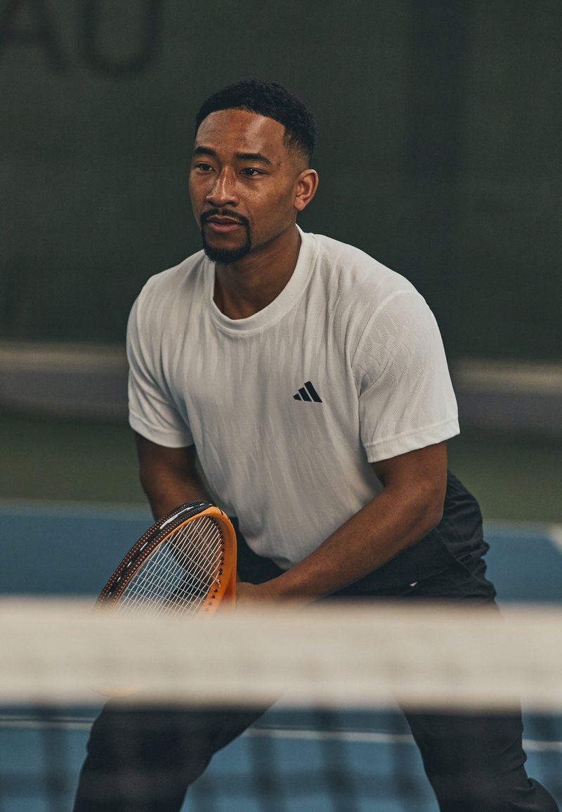 Sportlik mees valges tekstuuriga Adidas särgis ja mustades lühikestes pükstes, hoides oranž tennisereketit, paigutatud tennisemänguväljakule.