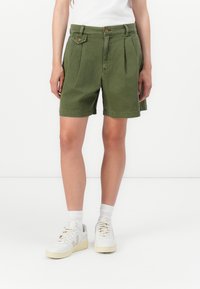 Calções verdes de algodão com pregas, cintura justa, dois bolsos frontais e detalhes laterais, combinados com ténis brancos e meias aos tornozelos.