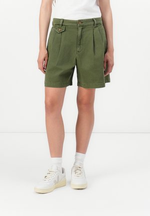 Pantaloni corti in cotone verde con pieghe, vita aderente, due tasche frontali e dettagli laterali, abbinati a scarpe da ginnastica bianche e calzini alla caviglia.