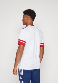 Weißes Sportshirt mit roten gestreiften Ärmeln und Akzenten, rundem Ausschnitt und glatter Textur, getragen mit dunkelblauen Hosen.