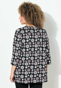 Femme aux cheveux bouclés portant un shirt à manches longues noir à motifs de flocons de neige blancs et rouges, se tenant contre un fond blanc.