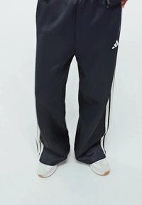 Pantalones deportivos negros hechos de un material suave con rayas laterales blancas. Tienen una cinturilla elástica y se complementan con zapatillas blancas.