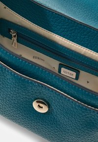 Torba na ramię z tealowej skóry z teksturowaną powierzchnią, zaokrąglonym kształtem, kieszenią na zamek i metalowym zatrzaskiem. Wnętrze zawiera etykietę z logo.