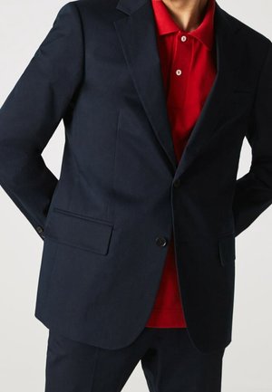 Marineblauwe op maat gemaakte colbert met twee voorzakken, één knoopsluiting en een gladde textuur, gedragen over een felrode poloshirt.