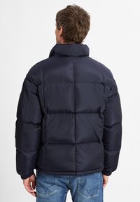 Giubbotto imbottito blu navy con colletto alto e design a baffle cucito. Presenta una texture liscia e lucida e polsini elasticizzati, abbinato a jeans blu.
