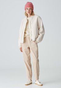 Veste bomber crème avec une finition texturée, pull en tricot crème épais, pantalon en velours côtelé beige clair et baskets blanches. Bonnet en tricot rose.