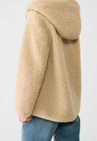 Beige tekstureret fleece hættetrøje båret over blå jeans, vist fra ryggen med en hånd synlig i siden.