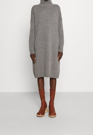 Vestido gris de punto con cuello alto hasta la rodilla, combinado con botas de cuero marrón hasta media pantorrilla con tacones de bloque de madera sobre un fondo liso.