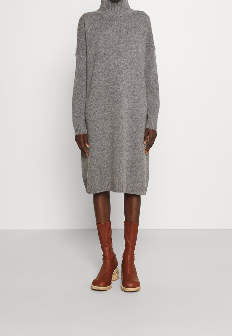 Robe en maille grise à col roulé longueur genou portée avec des bottes en cuir marron mi-mollet à talons blocs en bois sur fond uni.