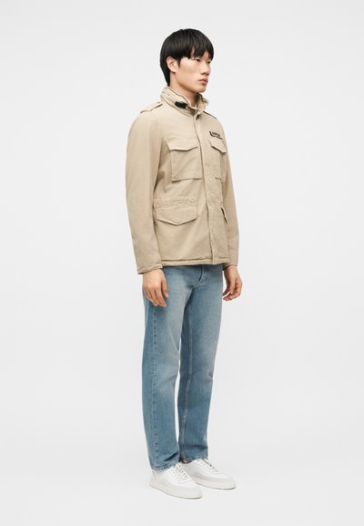 Jeune homme debout, portant une veste utilitaire beige avec poches avant, un jean bleu clair et des baskets blanches, sur un fond uni.