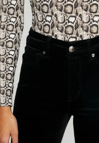 Femme portant un jean noir taille haute et un haut à manches longues avec un motif serpent beige et noir.
