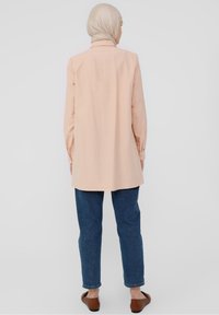 Camicia a maniche lunghe di colore rosa chiaro con colletto, taglio dritto e dettagli con polsini abbottonati, abbinata a pantaloni in denim blu e scarpe marroni.