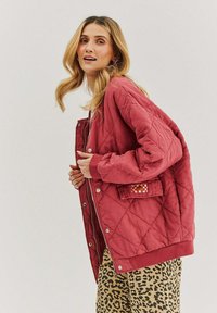 Prešívaná červená bunda s cvokcami a rebrovanými manžetami, so malou vzorovanou náprsní taškou; skombinovaná s nohavicami s leopardím vzorom.