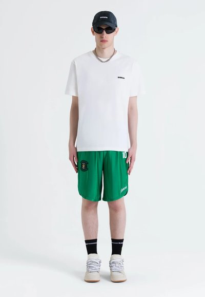 Biela tričko s čiernym logom, zelené šortky s emblémom, vrstvené ponožky a off-white tenisky. Príslušenstvo zahŕňa námornícku čiapku a čierne slnečné okuliare.