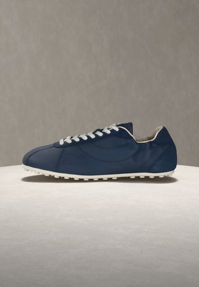 Ένα μόνο casual αθλητικό παπούτσι σε ναυτικό μπλε χρώμα με λευκά κορδόνια και σόλα βρίσκεται σε ανοιχτόχρωμη επιφάνεια με φόντο γκρι.