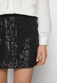 Mini-jupe noire à sequins avec des rayures verticales, mettant en avant des sequins circulaires brillants et une texture lisse. Une main est visible tenant la taille.