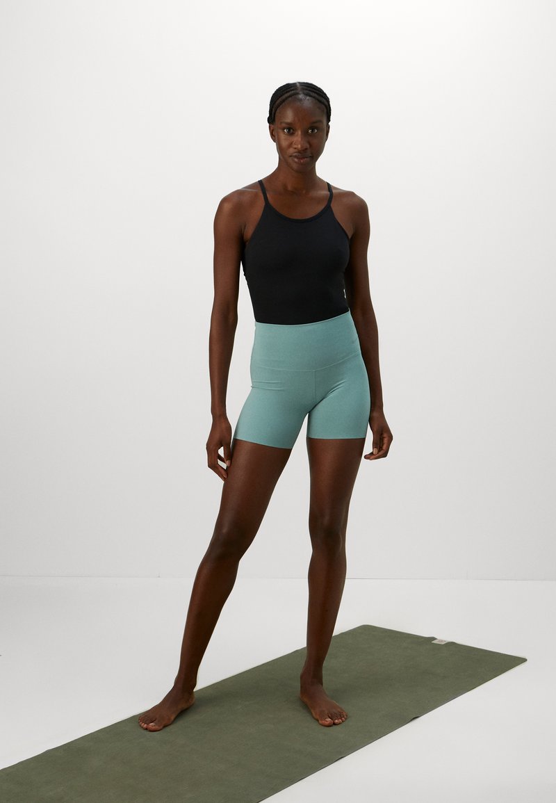 Zwarte tanktop en lichtblauwe high-waisted fietsshorts op een model, staand op een groene yogamat tegen een eenvoudige witte achtergrond.