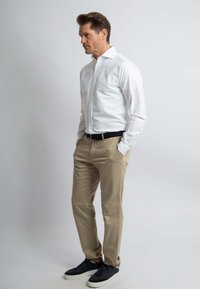 Witte button-up blouse, beige chinos, zwarte riem en zwarte sneakers; staande in profiel tegen een effen achtergrond.