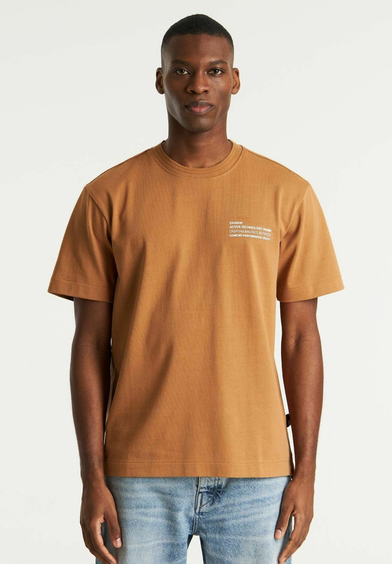 Brun bomulds T-shirt med korte ærmer og rund halsudskæring. Har hvid tekst på venstre front. Bæres med lyseblå denim.