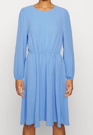 Vrouw draagt een lichtblauwe jurk met lange mouwen, een geplooide taille en een vloeiende knielange rok tegen een effen achtergrond.