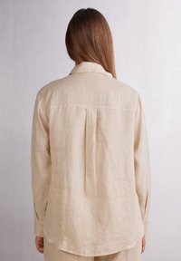 Camicia di lino beige con maniche lunghe e orlo arrotondato. Presenta una pence sul retro e una texture sottile. Design del colletto semplice e classico.