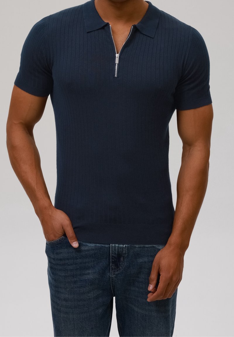 Homme portant un polo ajusté côtelé bleu marine à manches courtes avec fermeture éclair et un jean bleu foncé, debout avec une main dans la poche.
