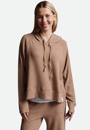 Lächelnde Frau mit blonden Haaren, die einen beigen Kapuzenpullover mit Kordelzug und passende Hose vor einem weißen Hintergrund trägt.