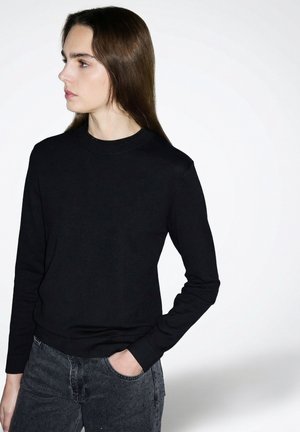 Jonge vrouw met lang bruin haar, gekleed in een zwart shirt met lange mouwen en donkergrijze jeans, kijkt naar links met één hand in haar zak.