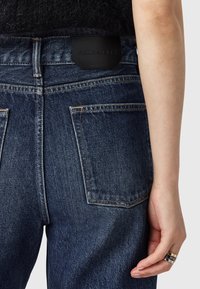 Jeans in denim blu scuro con finitura strutturata, caratterizzati da una patch in pelle visibile con logo "ALLSAINTS" in rilievo e dettagli di cucitura a contrasto.
