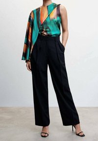 Buntes, asymmetrisches Oberteil in Grün mit orangefarbenen und schwarzen Mustern, kombiniert mit hochtaillierten, geraden schwarzen Hosen und schwarzen High Heels.