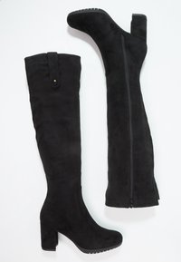 Botas negras de ante por encima de la rodilla con un tacón ancho, que cuentan con una cremallera lateral y una correa decorativa en la parte superior. Textura suave.
