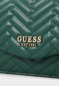 Teal quiltet læderpung med et guld "GUESS EST 1981" logo. Struktureret overflade med synlig syning i kontrasterende farver.
