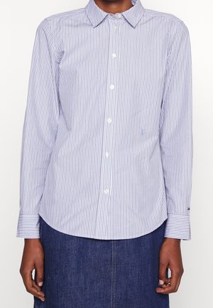 Personne portant une chemise blanche à fines rayures bleu clair à manches longues, avec un col, et une jupe en denim bleu foncé.