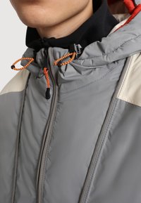 Grå multifunktionell jacka med hög krage, utrustad med orange dragband, svarta accenter och slät tygtextur. Dragkedjestängning fram.