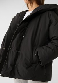 Schwarze Steppjacke mit Kapuze, mit durchgehender Knopfleiste, oversized Passform und strukturiertem Stoff. Inklusive Seitentaschen und Druckknöpfen.