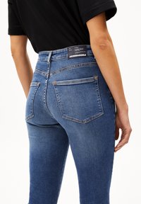Person wearing high-waisted blue denim jeans with back pockets and a black label on the waistband, paired with a black shirt.